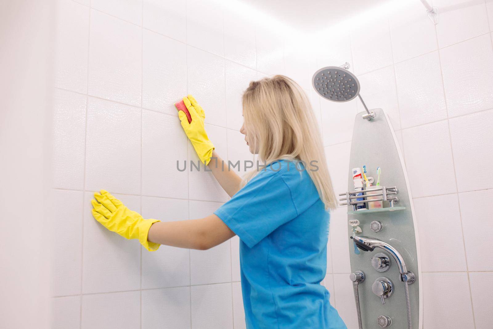 Blonde woman, employee cleaning company, wearing in yellow protective rubber gloves, with special cleaning sponge, cleaning wall tiled surface in white bathroom. Housework and housekeeping concept.