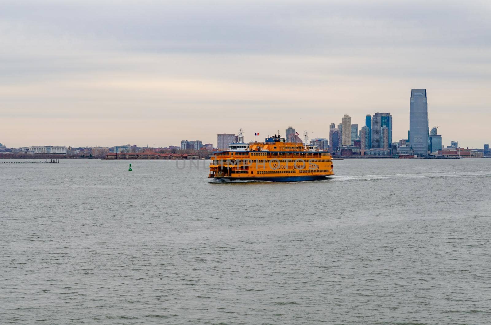 Yellow Staten Island Ferry on Hudson river passing by in front of Jersey City with office building skyscraper, New York, during winter evening with overcast, horizontal