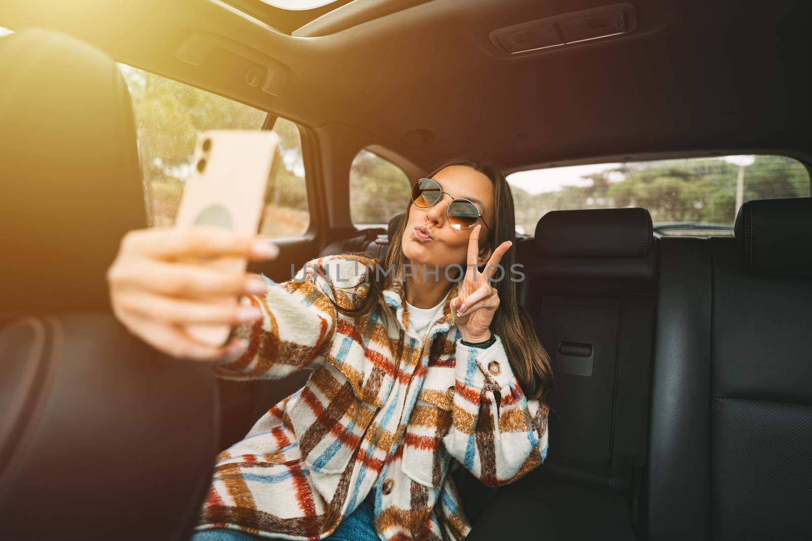 Stylish young woman sitting on back seat in the car and making selfie with smartphone. Woman taking picture with phone while sitting in taxi by DariaKulkova