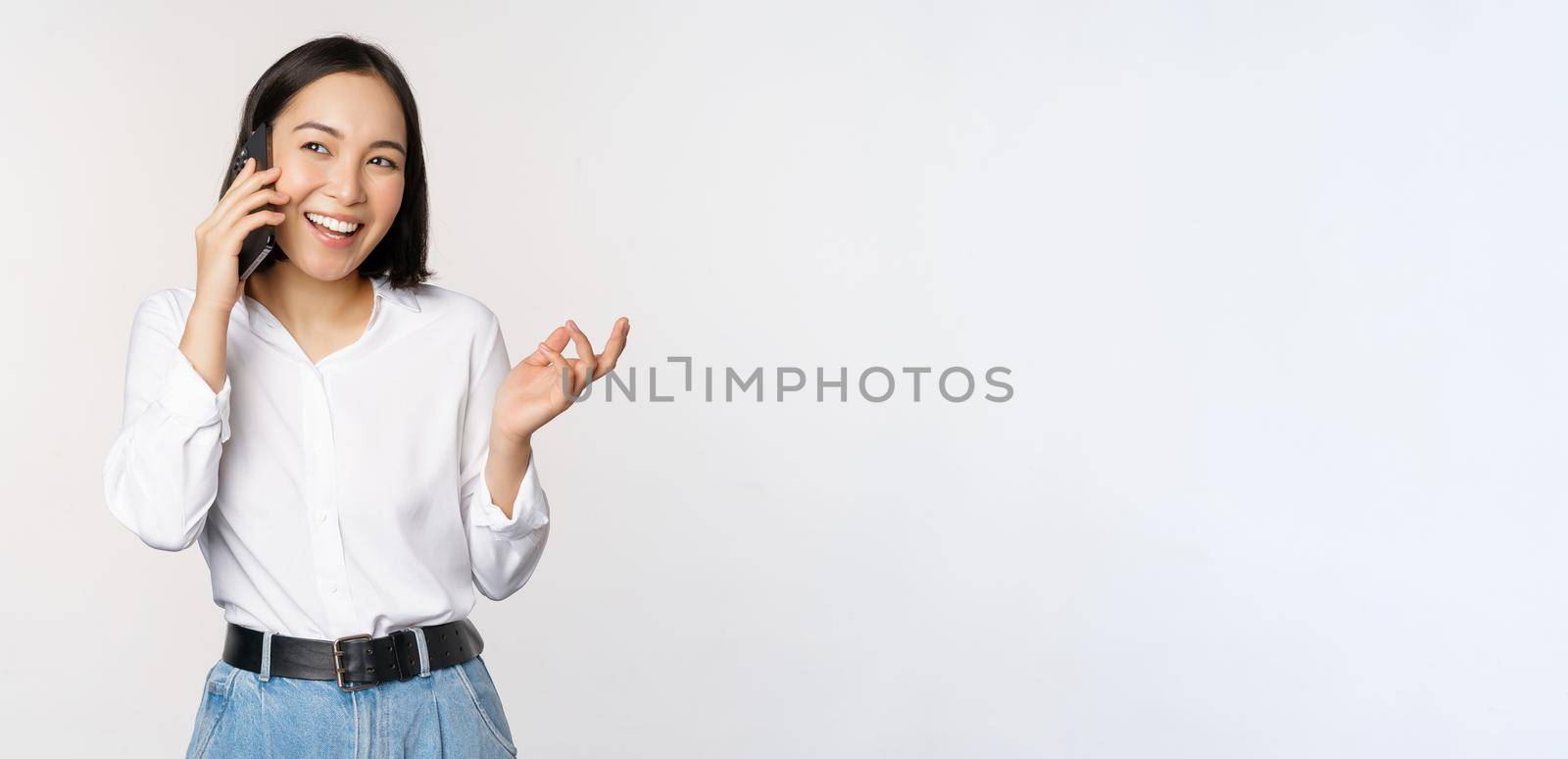 Friendly smiling asian woman talking on phone, girl on call, holding smartphone and laughing, speaking, standing over white background.