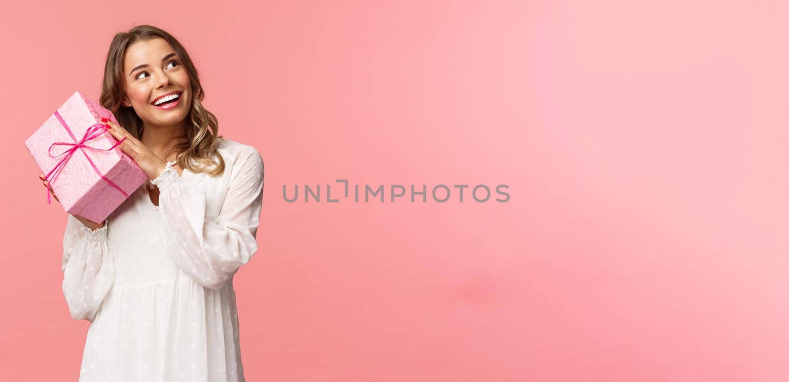 Holidays, celebration and women concept. Portrait of curious attractive young blond girl celebrating birthday, shaking pink gift box to found out whats inside, smiling intrigued look camera by Benzoix