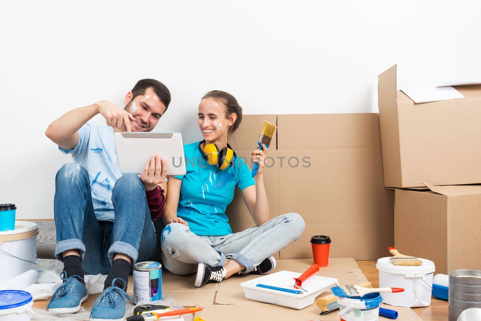 Happy couple watching something funny on tablet computer while sitting on floor. Painting tools and materials for home remodeling and interior redesign. Young family renovating their home after moving