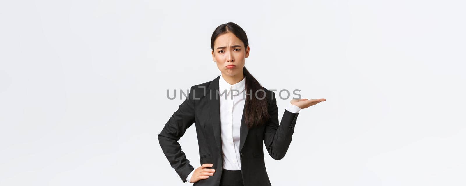 Disappointed and gloomy female sales manager, real estate agent in black suit having bad day, pouting and look upset while showing something, holding hand to the right, white background.