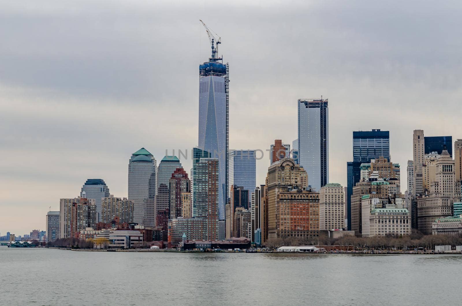 Skyline of Manhattan with One World Trade Center with construction area and Crane on top of it and Hudson River in forefront, during winter evening with overcast, horizontal