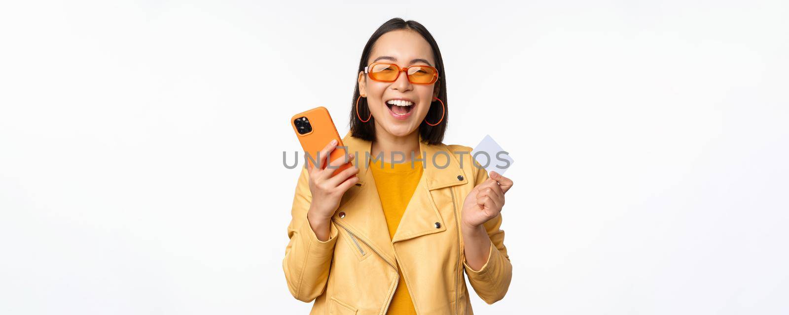 Online shopping and delivery concept. Happy korean girl in stylish clothes, holding credit card and smartphone, laughing and smiling, standing over white background.