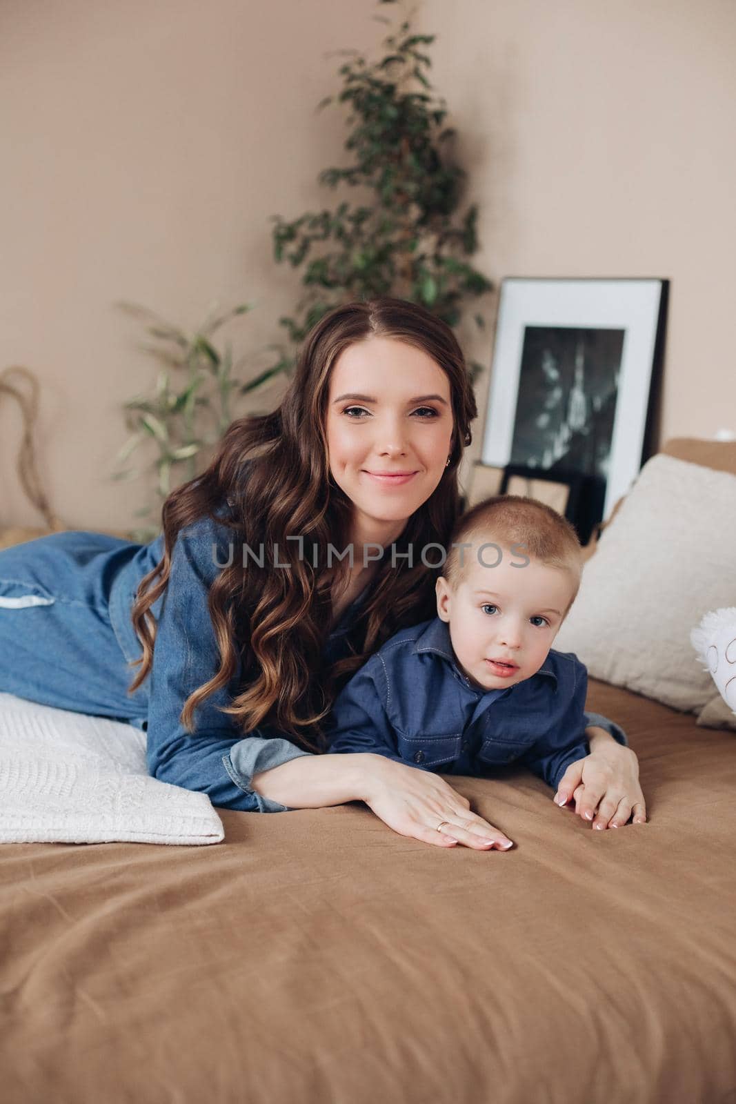 Portrait of gorgeous brunette young woman in casual denim dress embracing her lovely son in denim shirt on the bed. Family and motherhood concept.