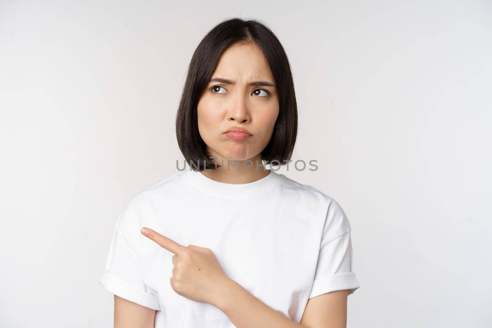 Angry and upset young asian woman pointing left, looking with regret or jealousy, feel unfair, standing over white background. Copy space