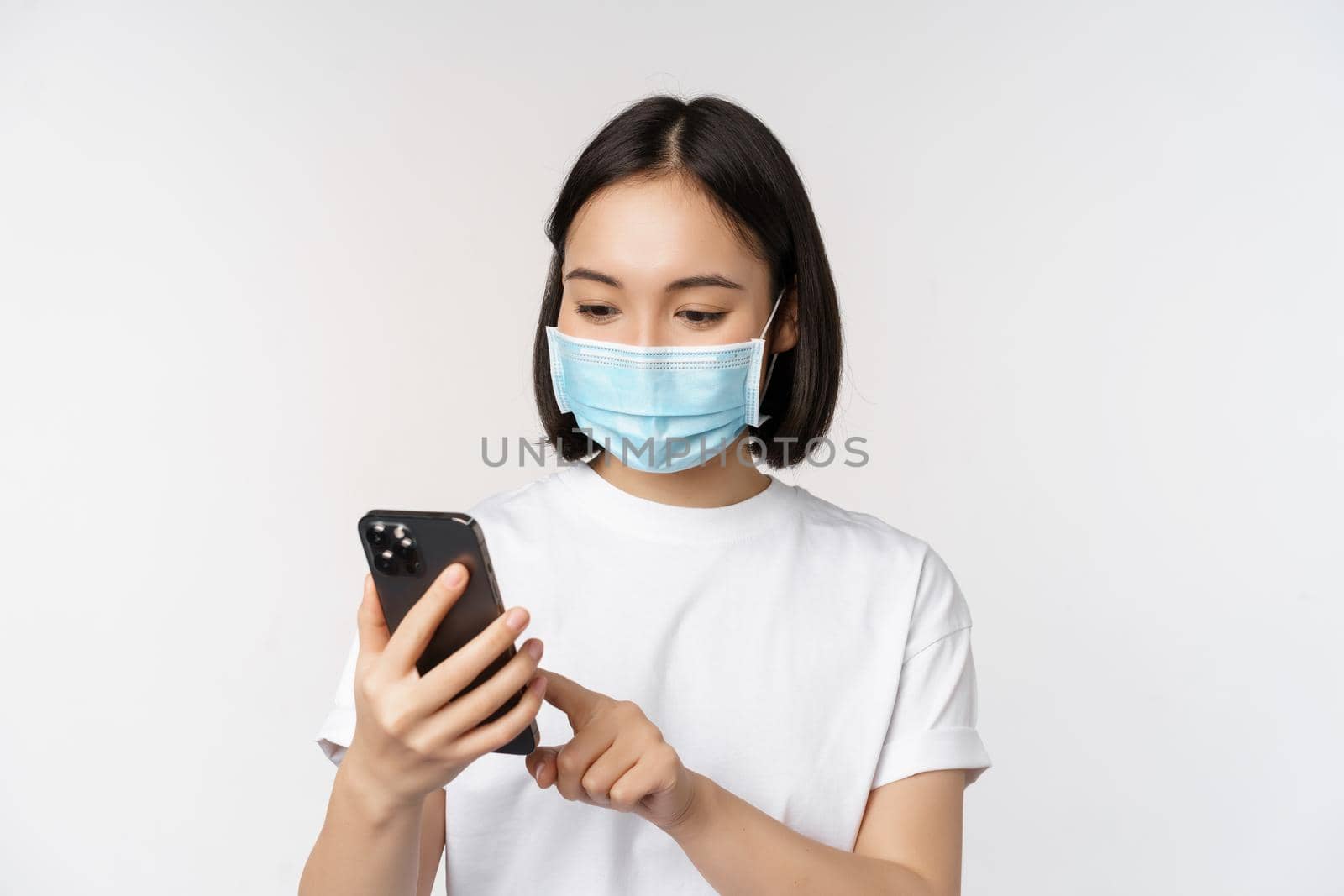 Health and covid-19 concept. Young asian woman in medical mask using mobile phone, typing on smartphone, standing against white background by Benzoix