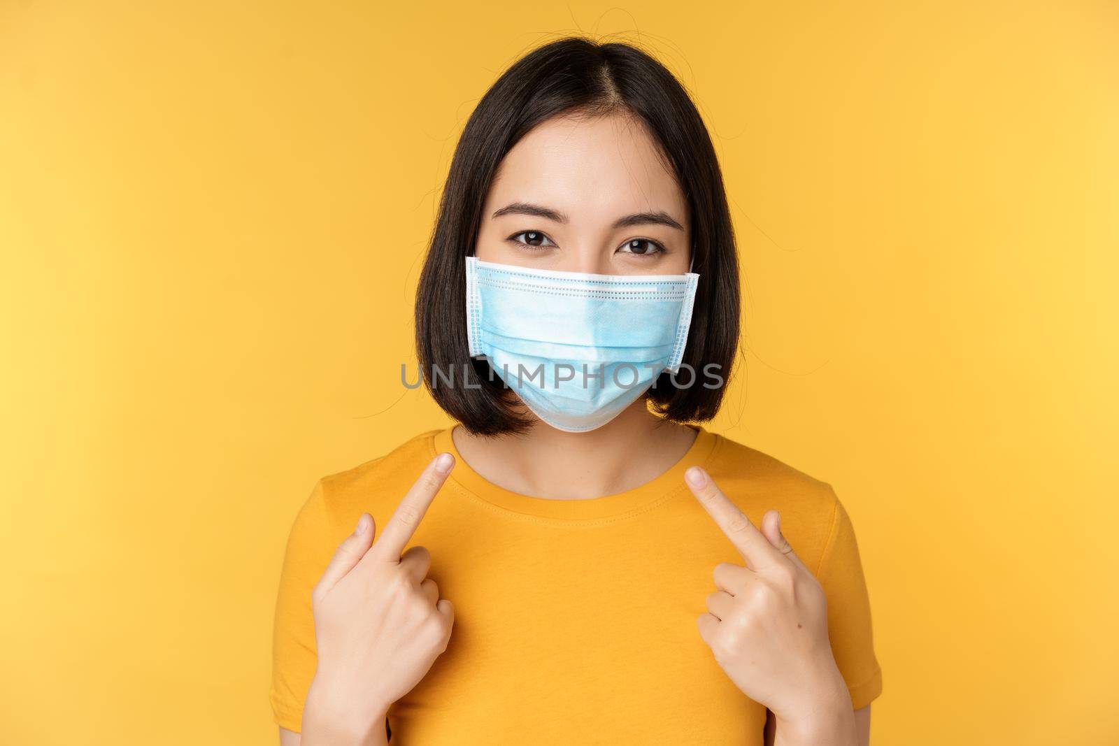 Portrait of smiling asian woman in medical face mask, pointing at her personal protective equipment from covid-19 during pandemic, standing against yellow background.