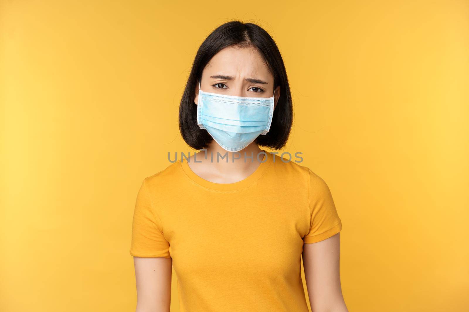 Image of sad and disappointed asian woman in medical mask, frowning and looking displeased, standing against yellow background by Benzoix