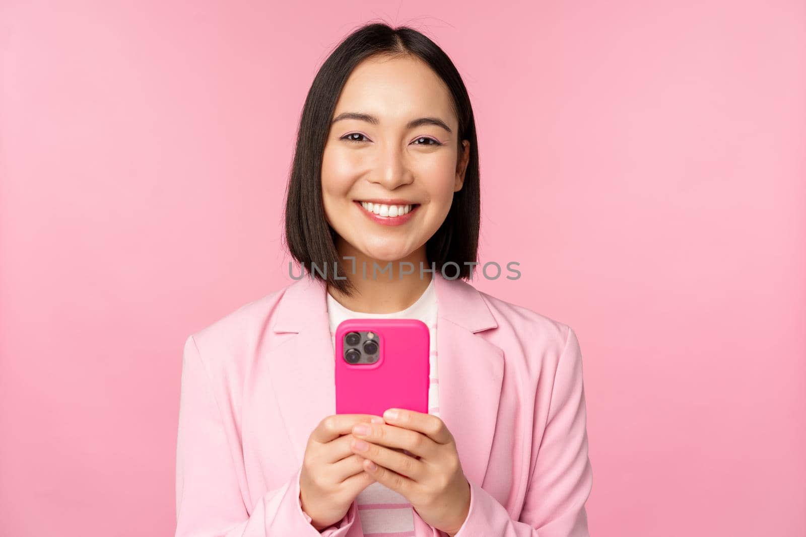 Portrait of smiling business woman, asian corporate person using smartphone, mobile phone application, standing over pink background.