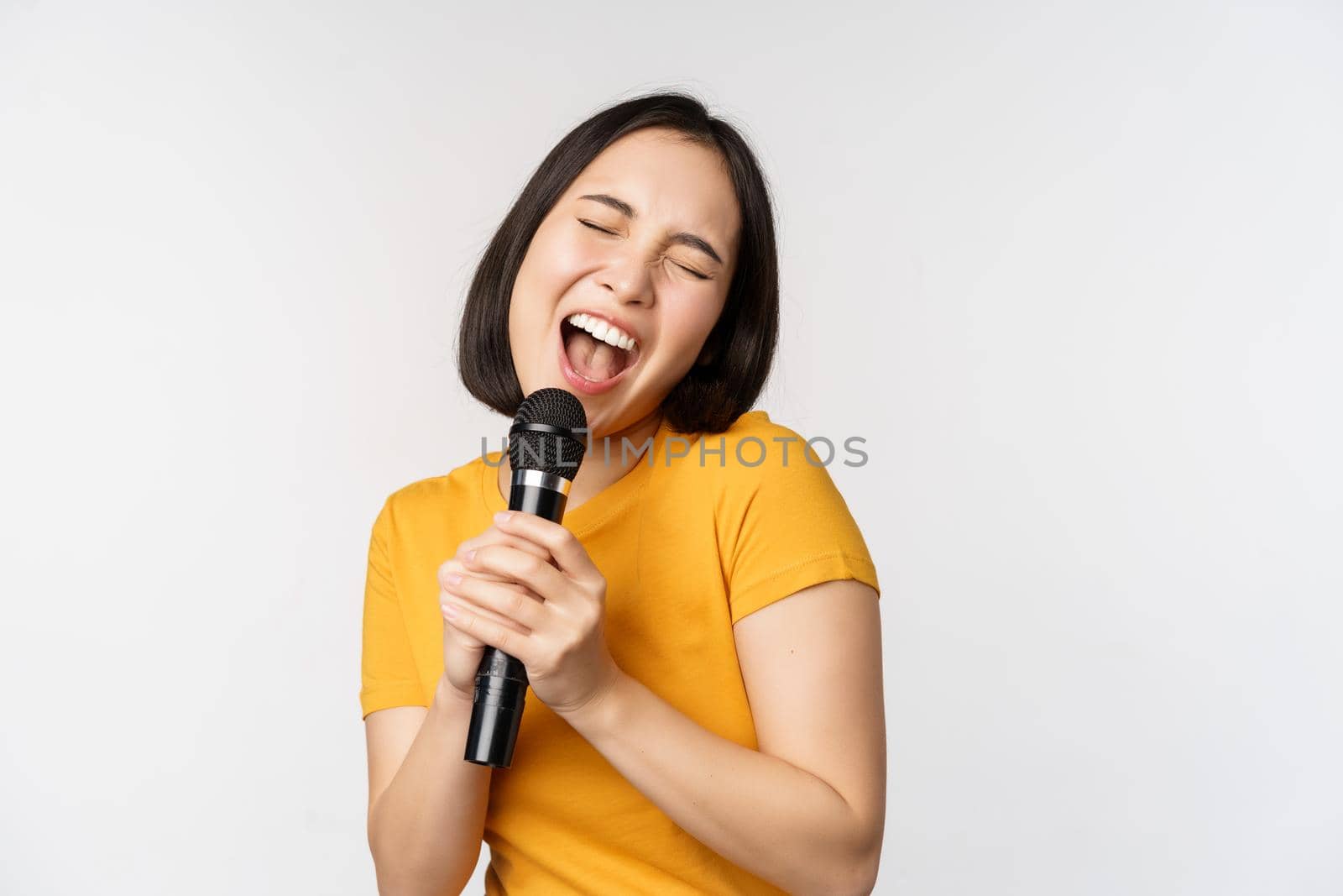 Happy asian girl dancing and singing karaoke, holding microphone in hand, having fun, standing over white background.