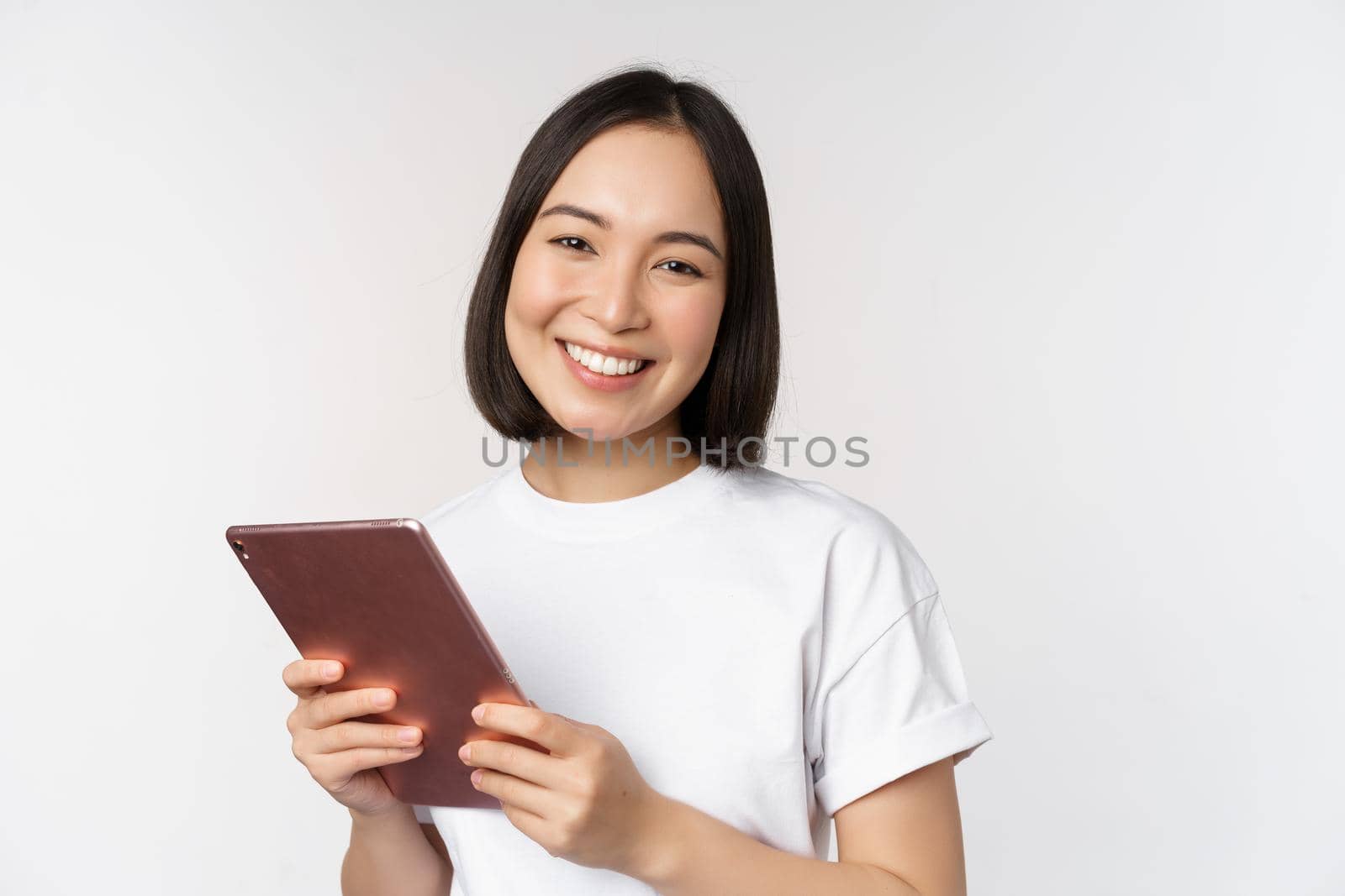 Smiling asian girl with digital tablet, looking happy and laughing, posing in tshirt over white background by Benzoix