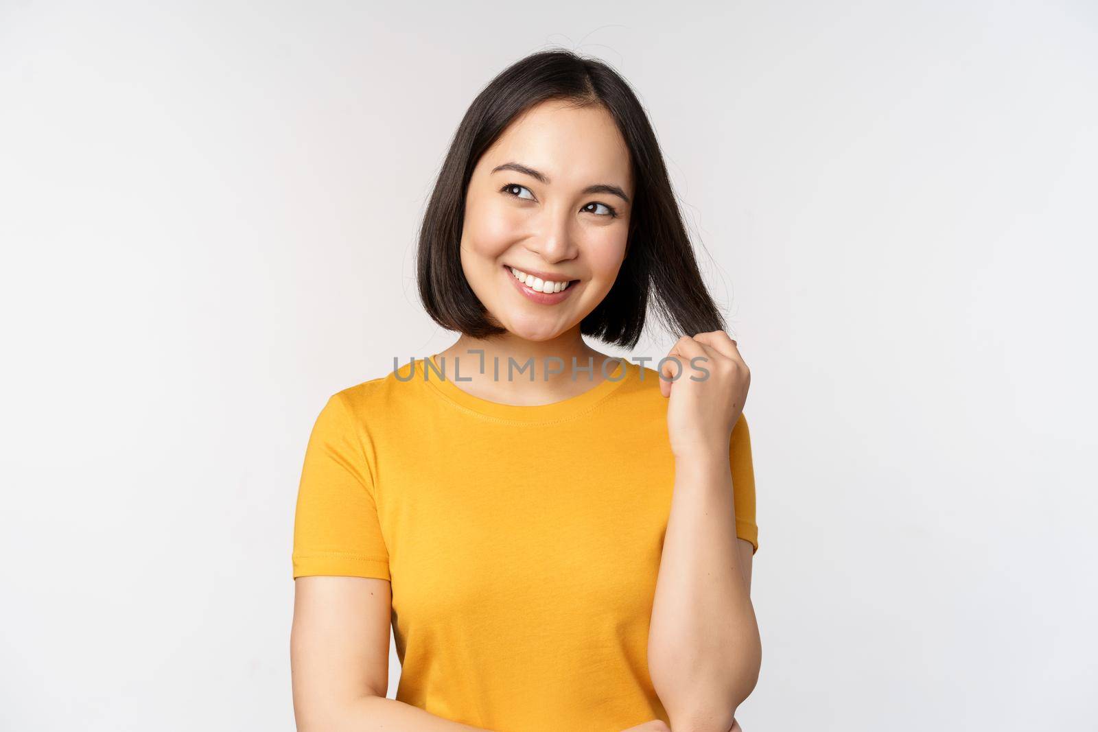 Beautiful romantic asian girl, smiling and playing with hair, looking happy at camera, standing in yellow t-shirt over white background.