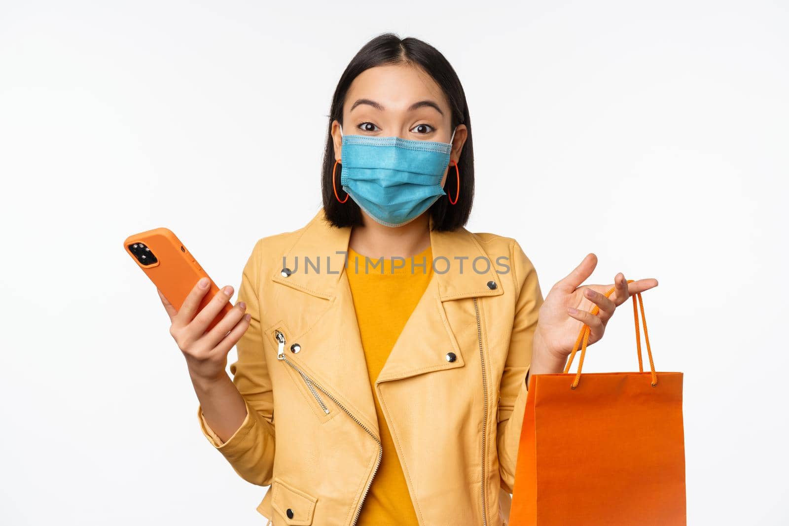 Image of stylish asian girl shopper, holding smartphone and shopping bag without store logo, wearing medical face mask from covid-19, white background.