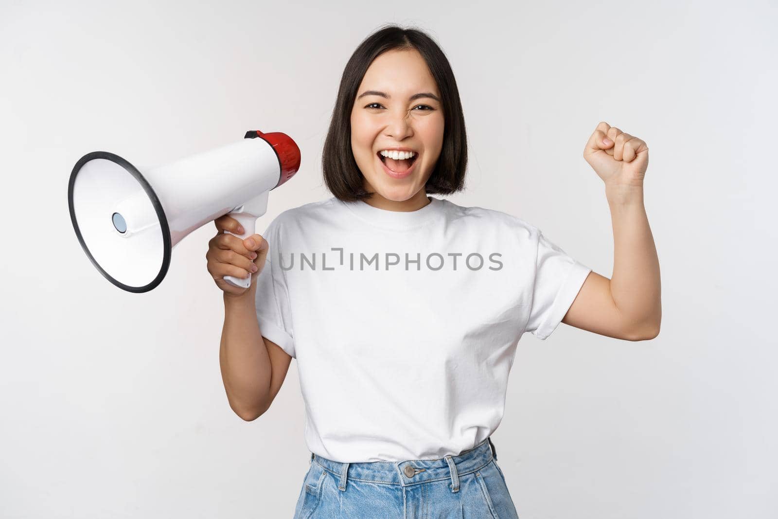 Happy asian woman shouting at megaphone, making announcement, advertising something, standing over white background by Benzoix