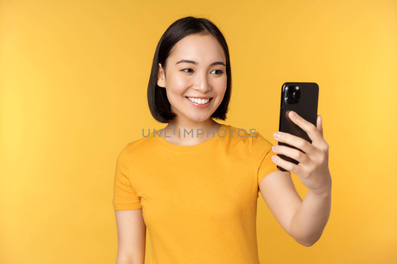 Image of happy, beautiful asian girl video chatting, talking on smartphone application, standing against yellow background. Copy space