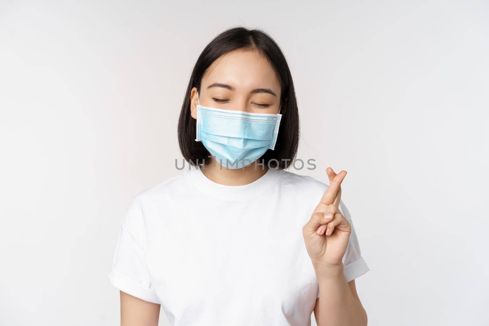 Covid-19, healthcare and medical concept. Image of asian girl in medical face mask, cross fingers, praying, making wish and smiling, standing over white background.
