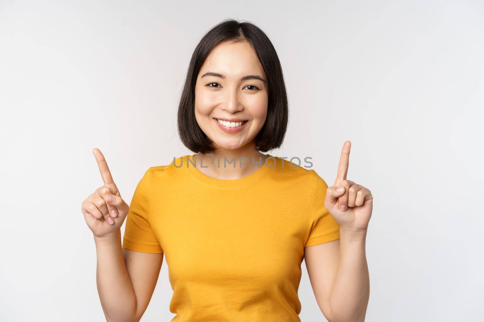 Portrait of beautiful japanese girl smiling, pointing fingers up, showing advertisement, standing in yellow tshirt against white background by Benzoix