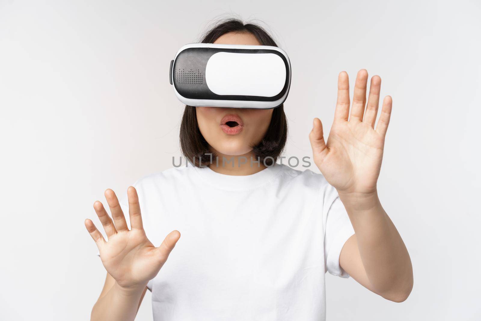 Amused asian girl using VR glasses, virtual reality headset and reaching hands into empty space, touching smth augmented, standing over white background.