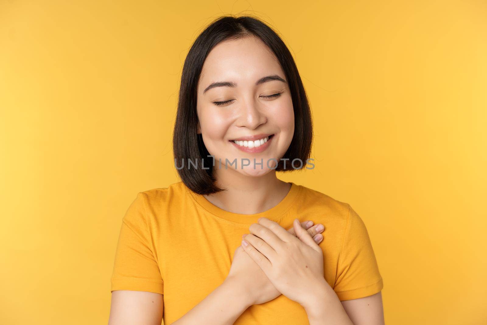 Beautiful asian woman, smiling with tenderness and care, holding hands on heart, standing in tshirt over yellow background.