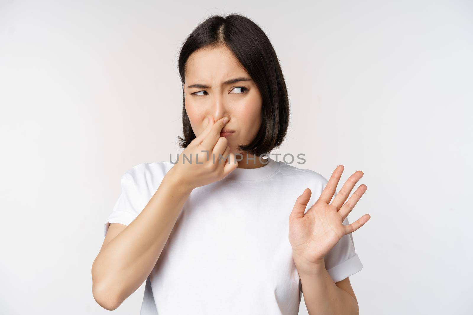 Asian girl looks disgusted, rejecting product with bad smell, shut nose from aversion and cringe, standing against white background by Benzoix
