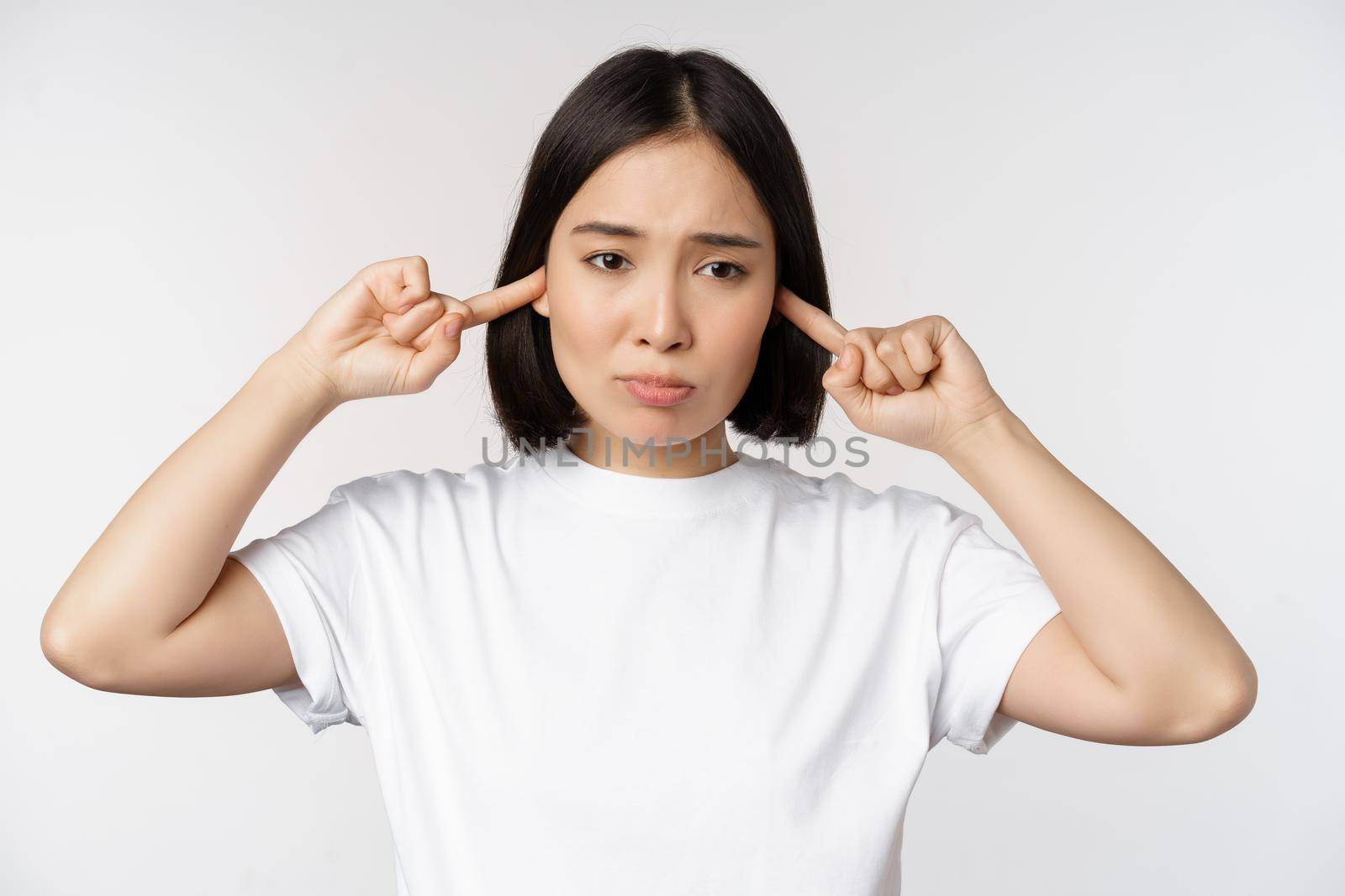 Portrait of asian woman shut ears and feeling discomfort from loud noise, annoying sound, standing over white background.