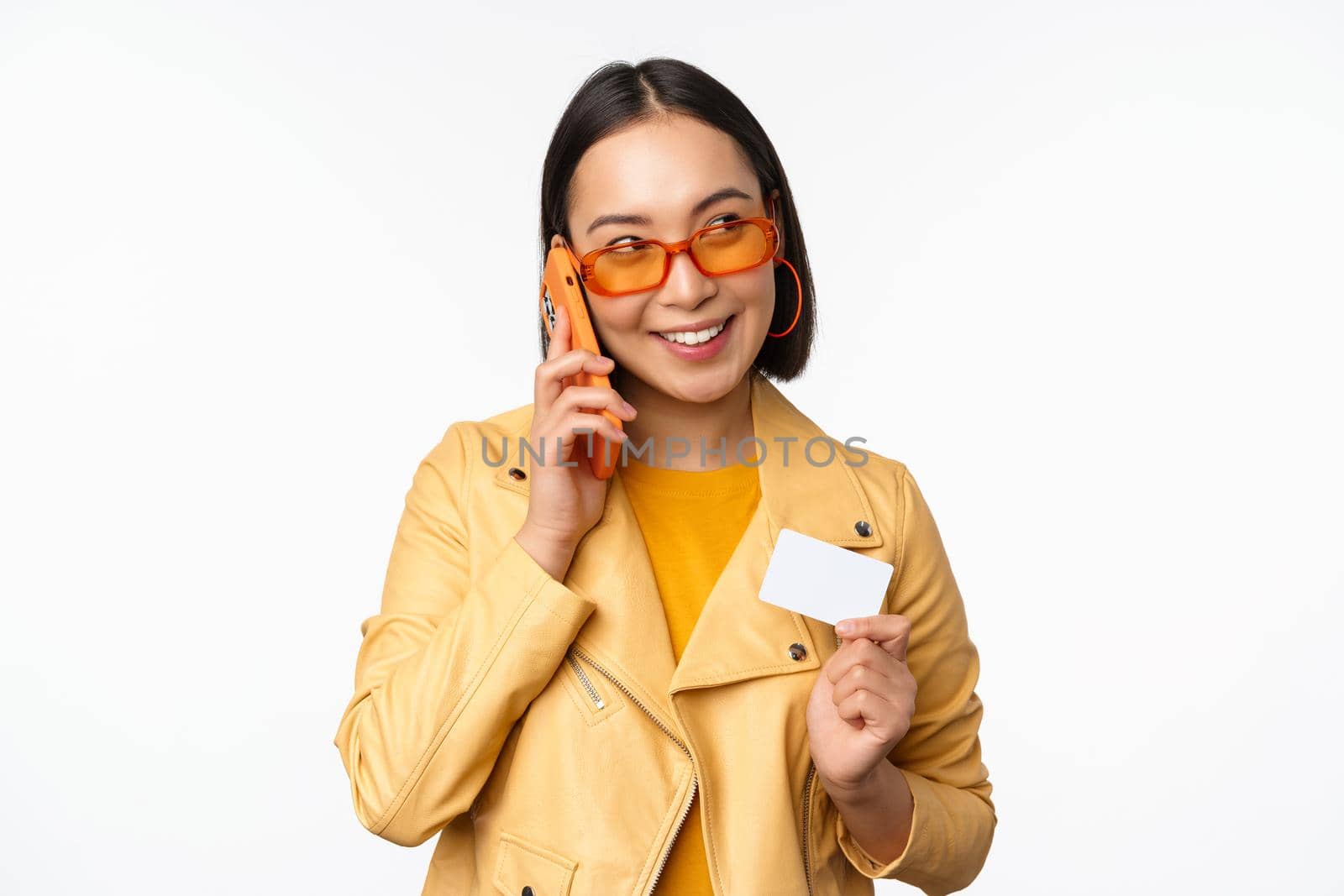 Stylish asian female model, talking on smartphone and showing credit card, standing over white background. Copy space