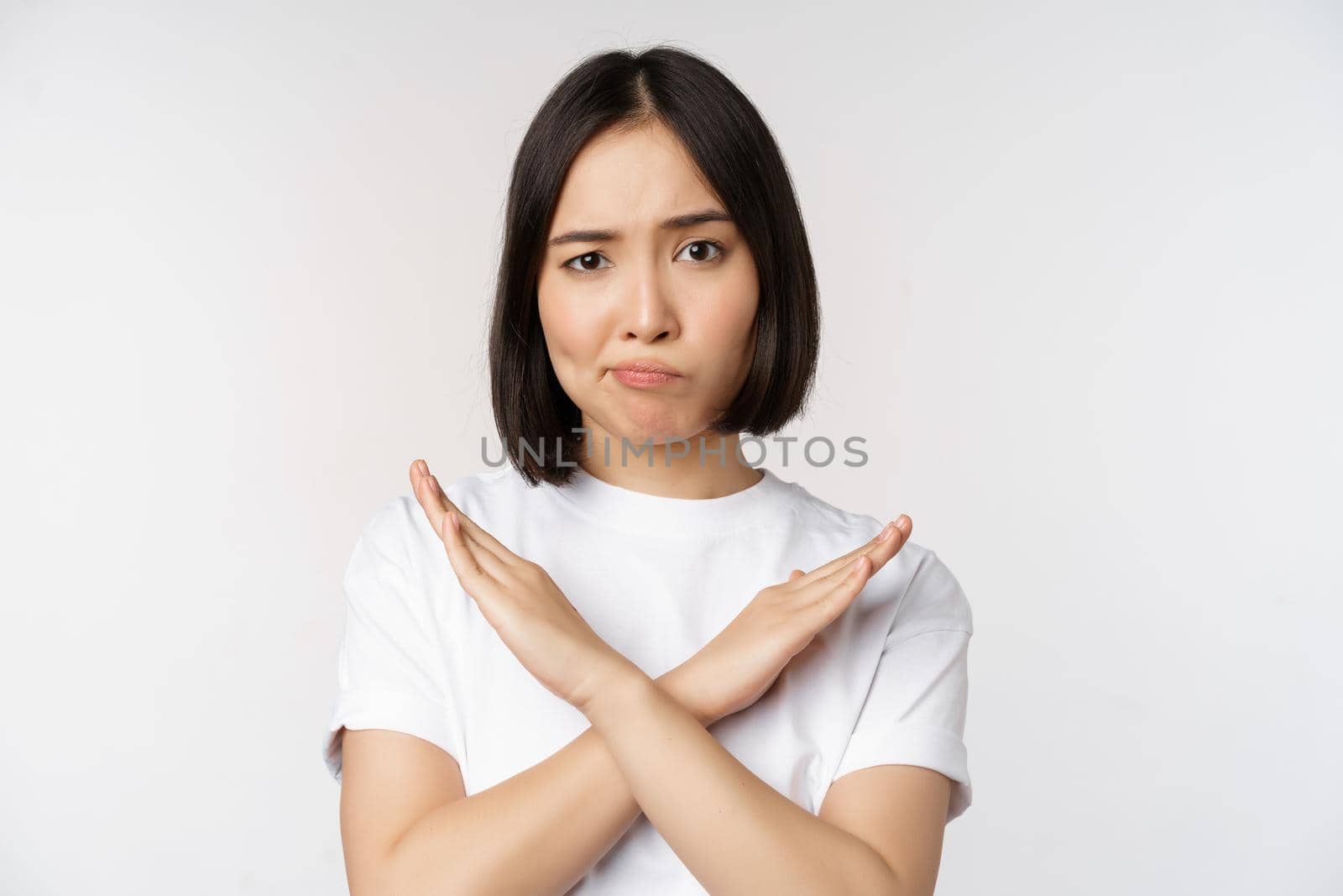 Portrait of asian korean woman showing stop, prohibition gesture, showing arm cross sign, standing in tshirt over white background by Benzoix