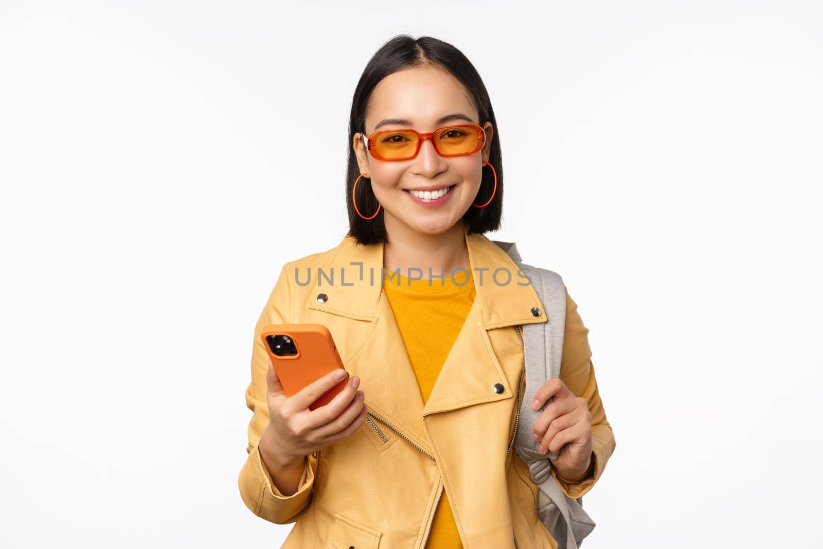 Stylish young asian woman tourist, traveller with backpack and smartphone smiling at camera, posing against white background. Copy space