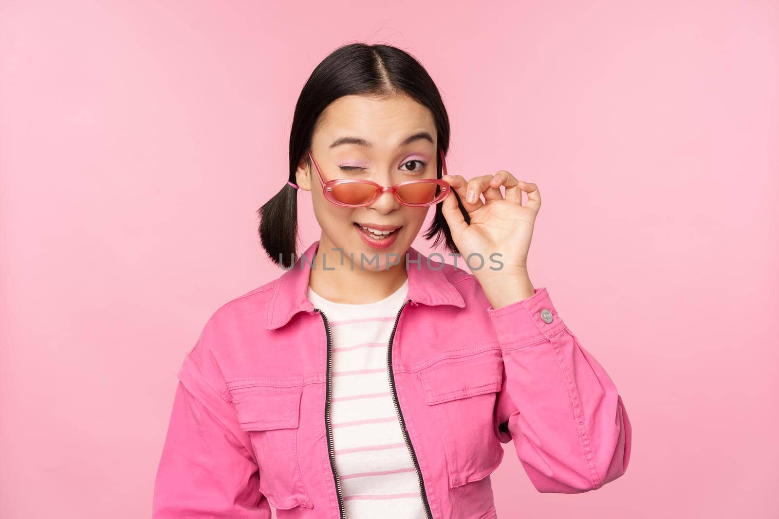 Eyewear advertisement. Stylish modern asian girl touches sunglasses, wears pink, poses against studio background. Copy space.