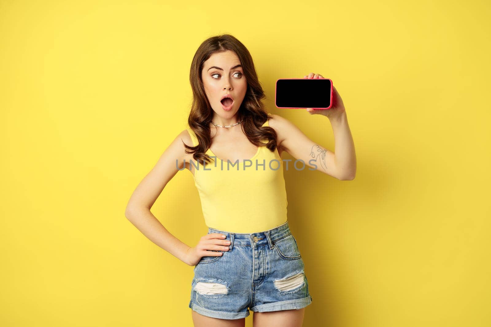 Shocked and amazed stylish girl, showing smartphone screen, app on horizontal mobile phone display, standing in summer clothes over yellow background by Benzoix