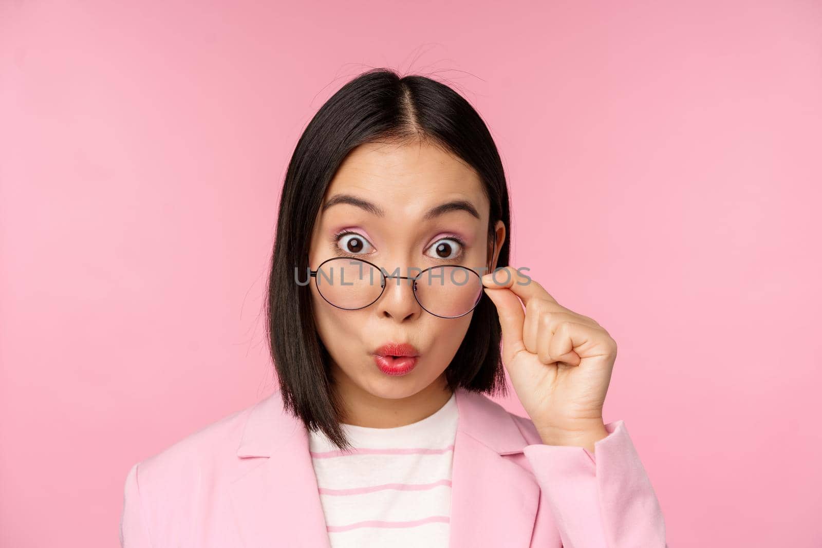 Close up portrait of korean office lady takes off glasses and looking impressed at camera, surprised face expression, pink background.