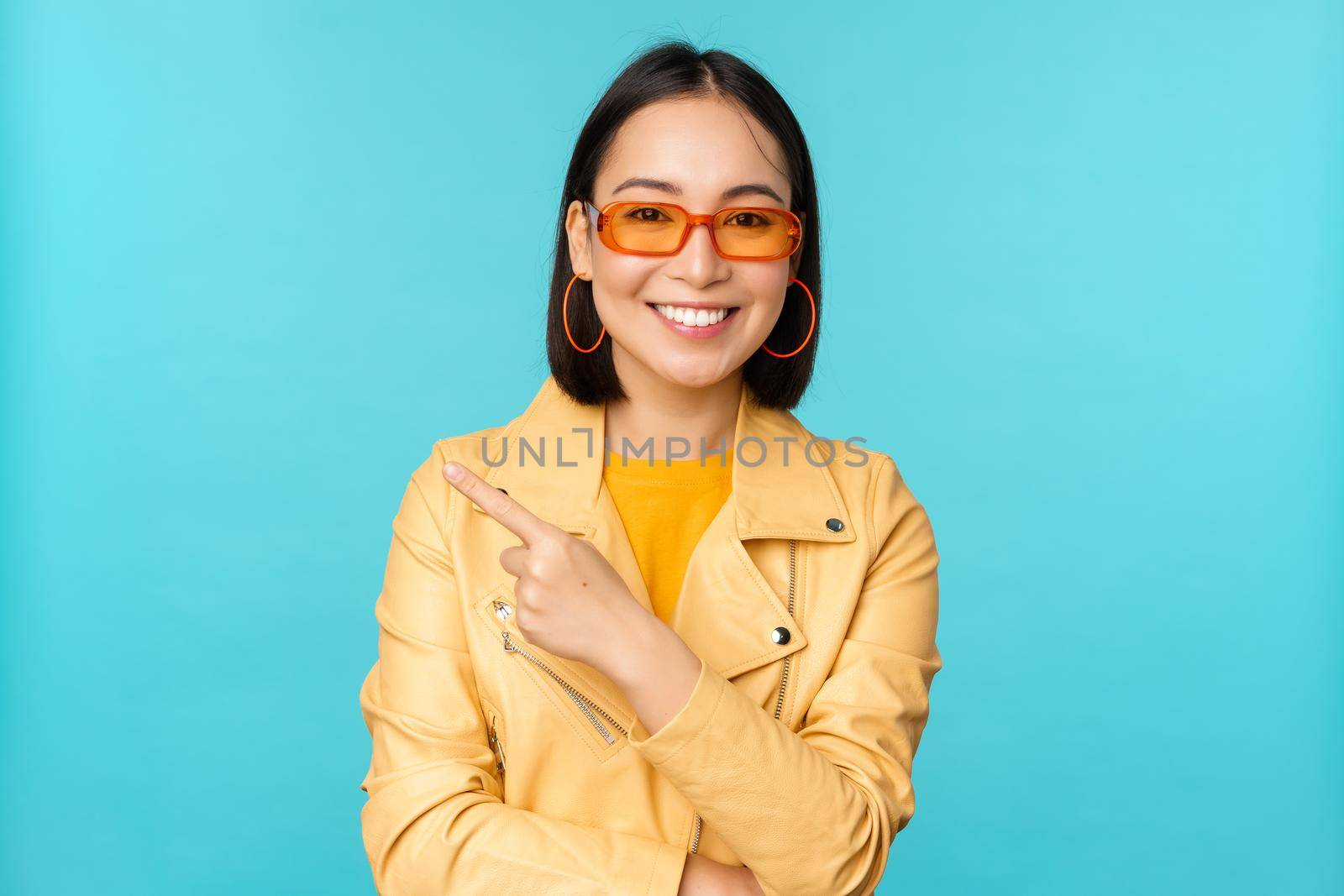 Stylish smiling asian woman in sunglasses, pointing finger left, showing advertisement banner, standing in trendy yellow coat over blue background.