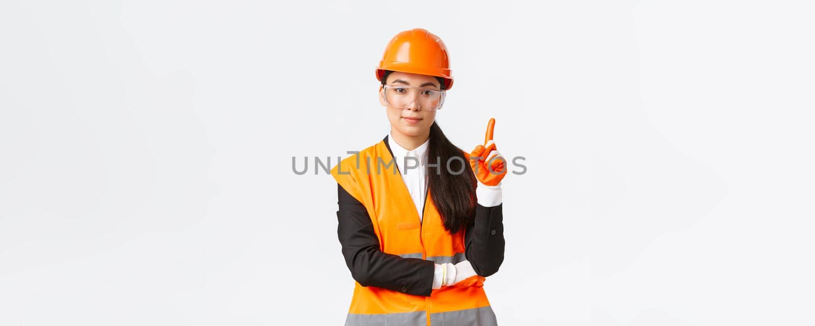 Safety first. Serious, smiling female asian construction engineer, industrial woman showing finger at telling rules on enterprise, wearing protective glasses, helmet and gloves, white background.