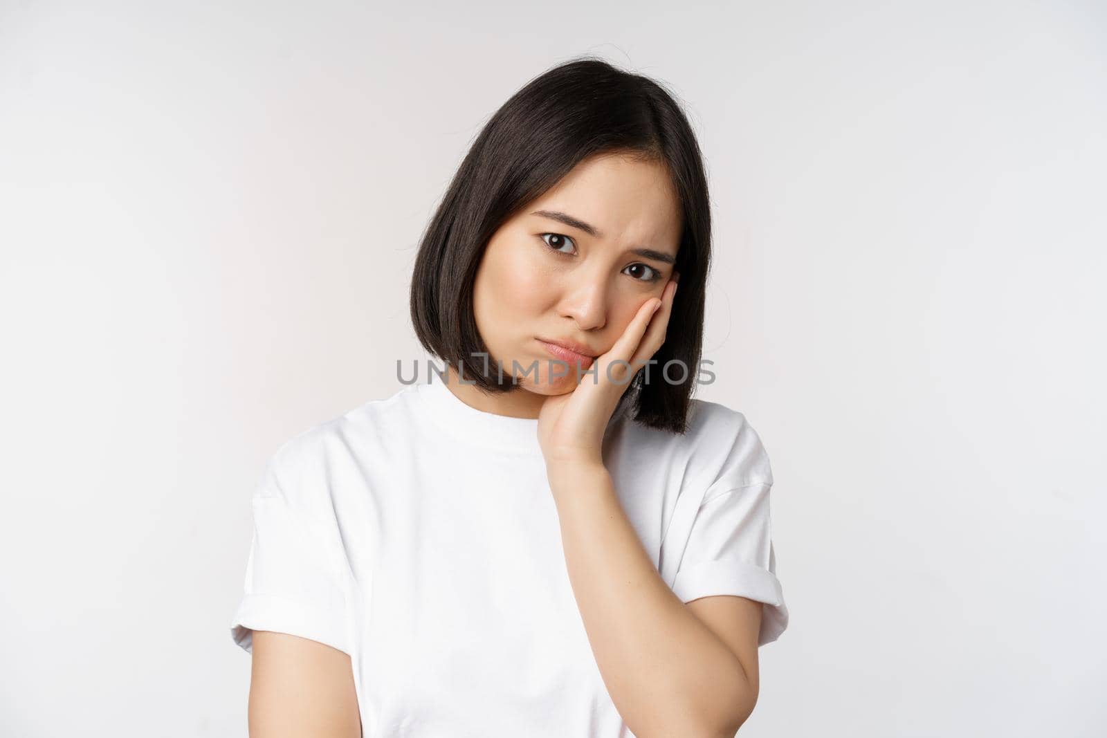 Sad asian girl looking upset and lonely, sulking and frowning, standing against white background in casual tshirt.