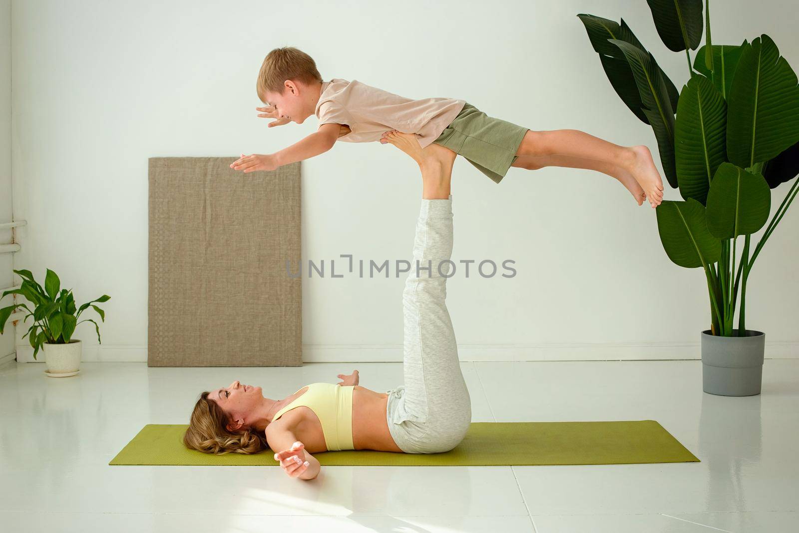 A woman, doing yoga with a child boy, lies on a sports mat, raising her legs up, doing exercises with support. by Zakharova