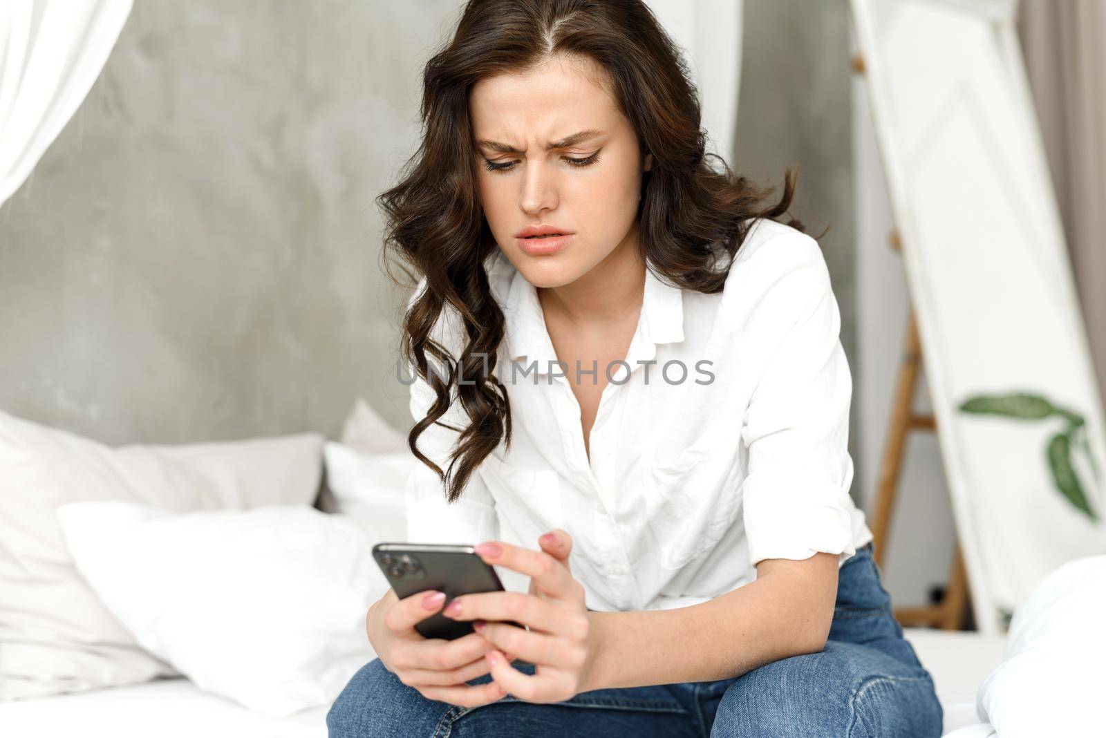 A young woman scrolls through her social media feed and sees some bad news. Caucasian female model sits on the bed and at home and worries, reading about problems, spam in the mail by etonastenka