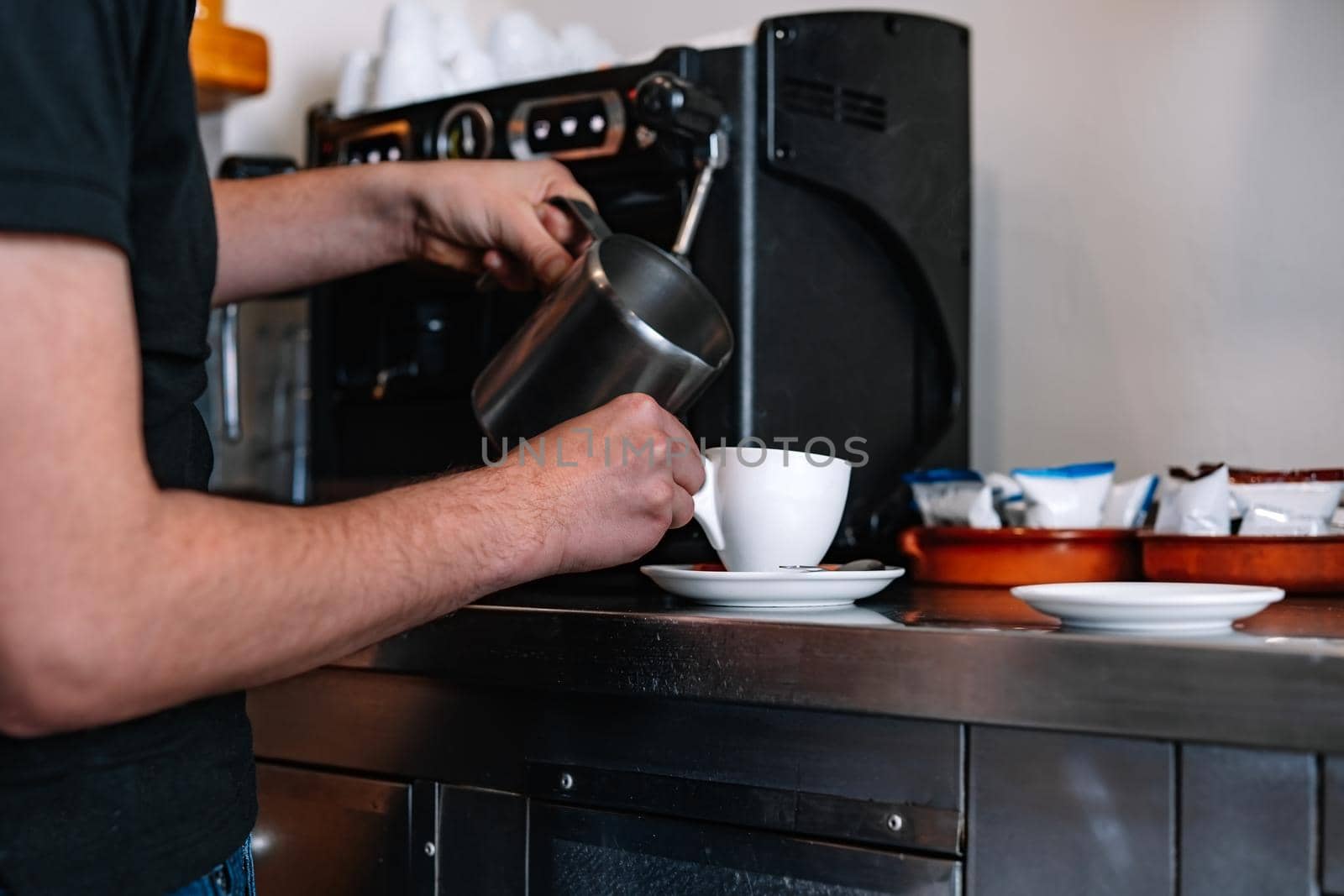 Young waiter with long hair tied back, concentrated and hard working, dressed in company uniform a black polo shirt, pouring milk into a cup of coffee. Restaurant. Preparing coffee with milk. Warm atmosphere and dim lighting. Steaming cups of coffee. Horizontal.