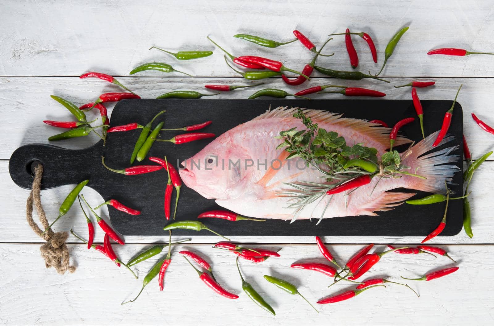 raw fresh pink tilapia fish lies on a board sprinkled with small red and green hot peppers, pink Himalayan salt and garnished with fresh curry sprigs, on white wooden planks. High quality photo