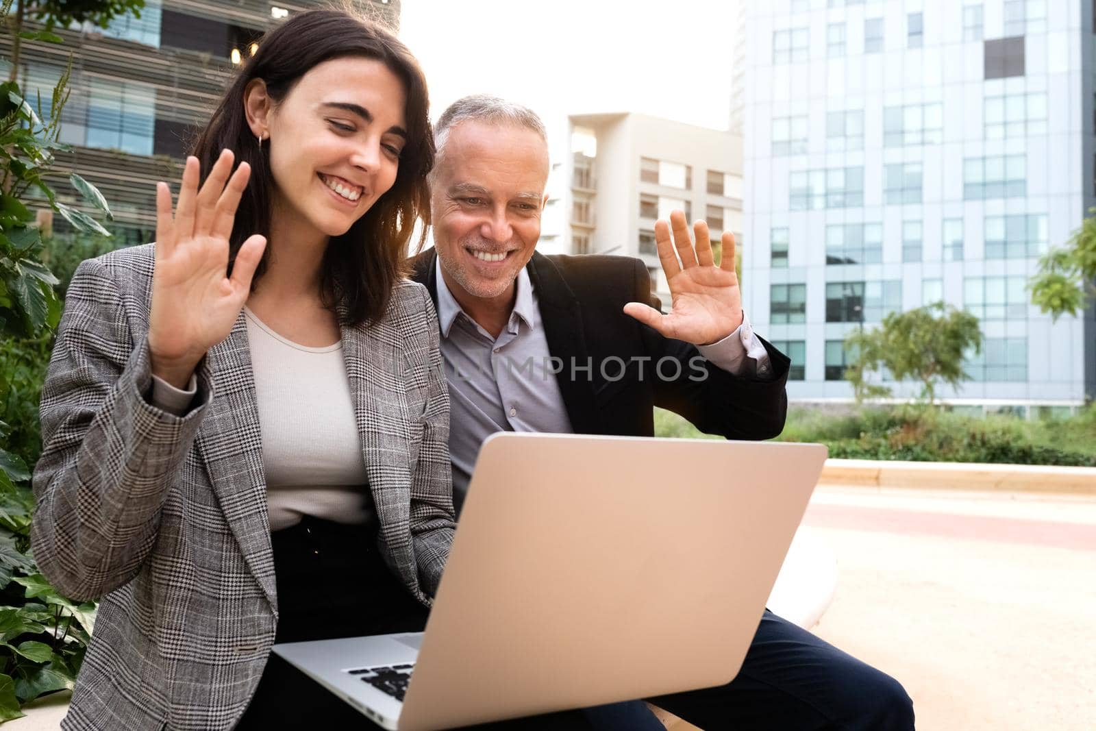Young woman and caucasian man wave hello on business video call sitting on a bench outside office buildings. Copy space. Technology concept.