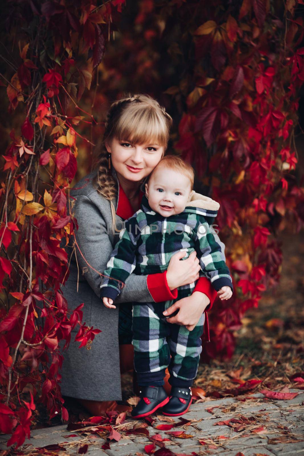 Professional portrait of attractive young woman with fair hair in braid embracing her little baby girl in plaid warm overall while standing in beautiful bright tree with red leaves. They are smiling at camera surrounded by vivid foliage in autumnal park.