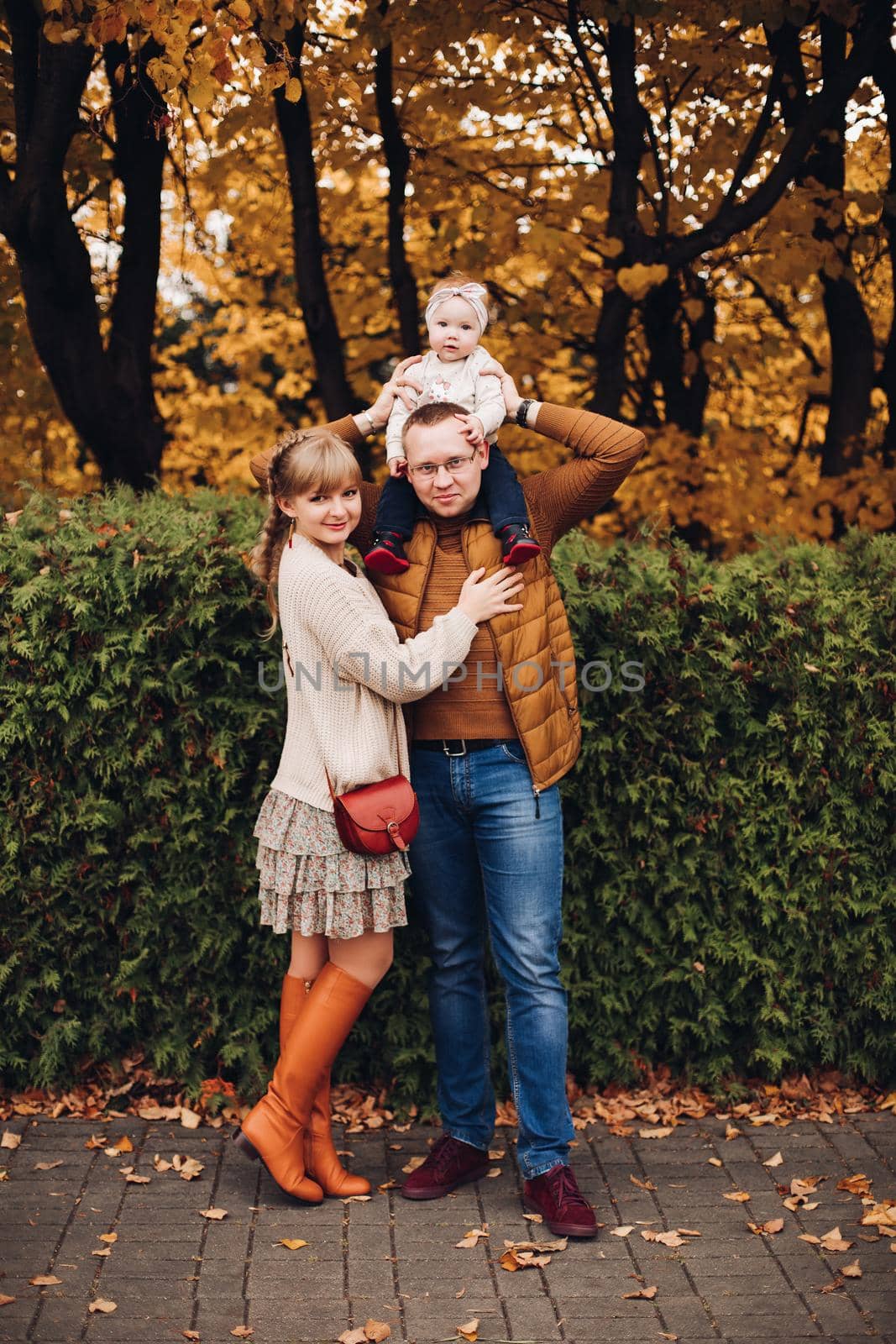 Portrait of attractive young mother and handsome smiling father wearing glasses holding their beautiful lovely baby girl on hands standing against green hedge in autumnal park. They are smiling and looking at camera.