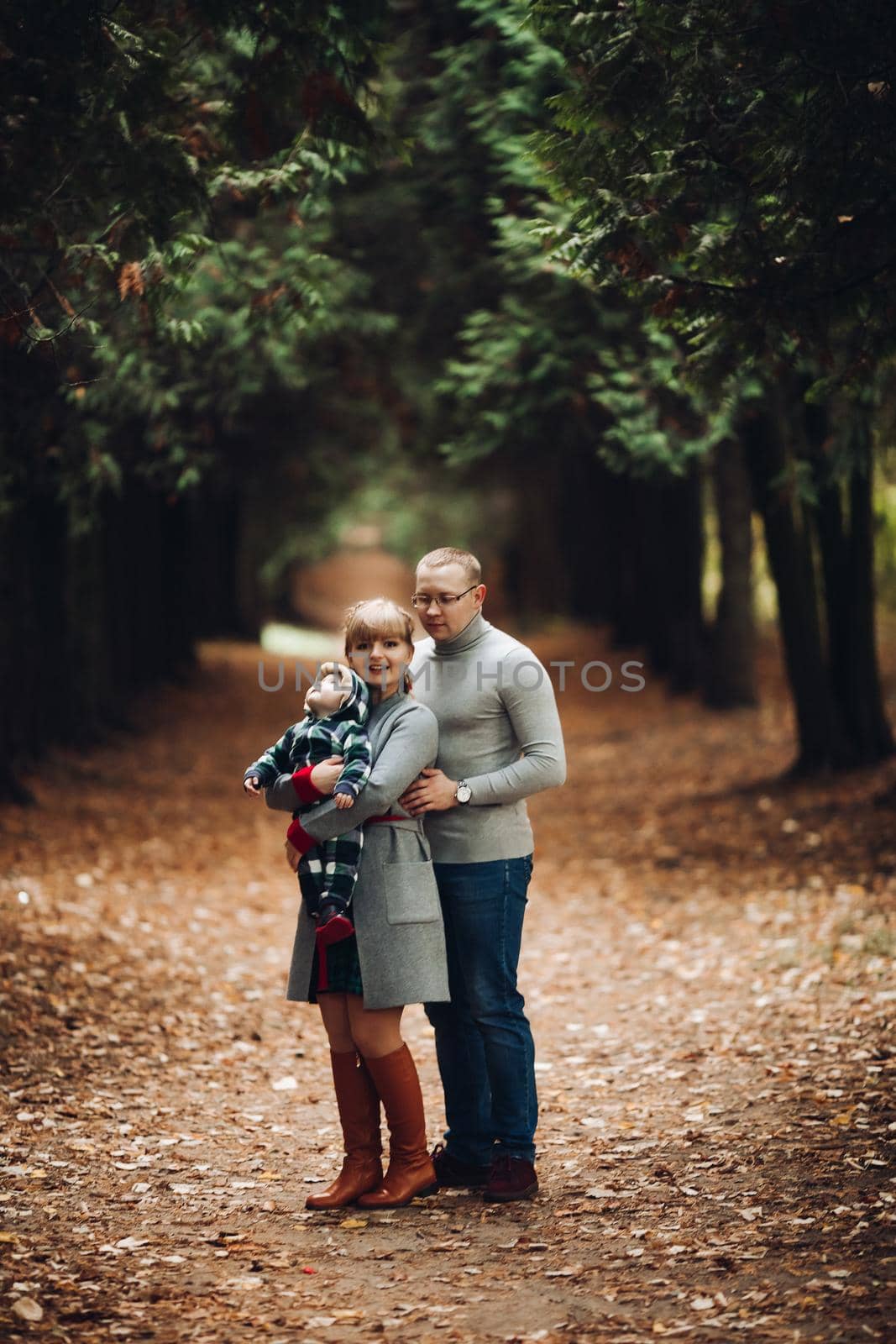 Portrait of attractive young mother and handsome smiling father wearing glasses holding their beautiful lovely baby girl on hands standing against green hedge in autumnal park. They are smiling and looking at camera.