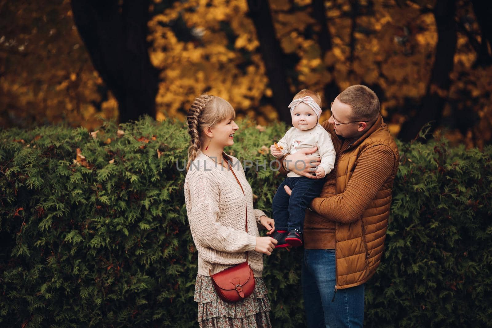 Portrait of attractive young mother and handsome smiling father wearing glasses holding their beautiful lovely baby girl on hands standing against green hedge in autumnal park. They are smiling and looking at camera.