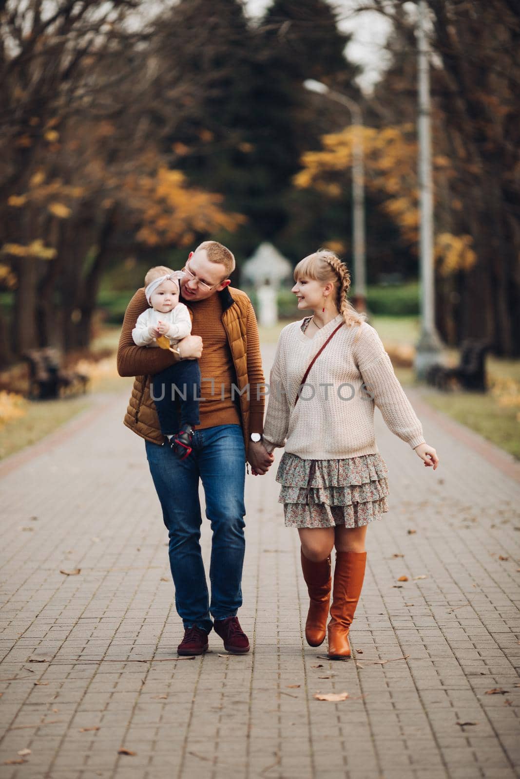 Portrait of attractive young mother and handsome smiling father wearing glasses holding their beautiful lovely baby girl on hands standing against green hedge in autumnal park. They are smiling and looking at camera.