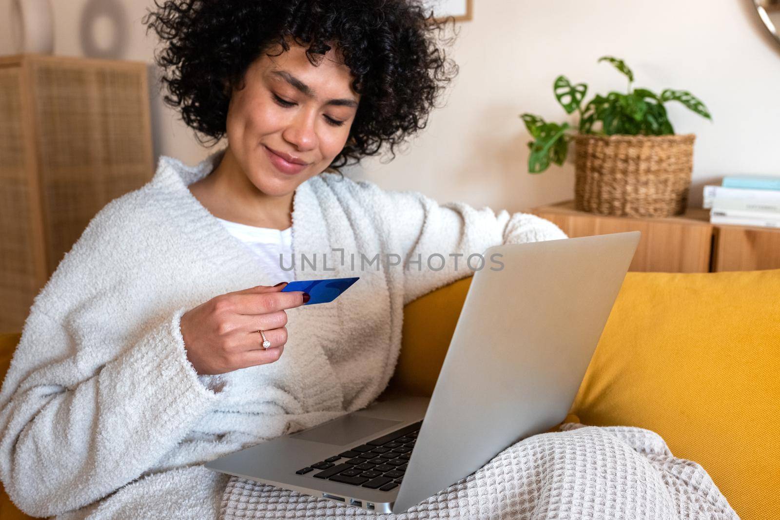 African american woman using laptop and reading credit card information to shop online at home sitting on sofa. Lifestyle and e-commerce concept.