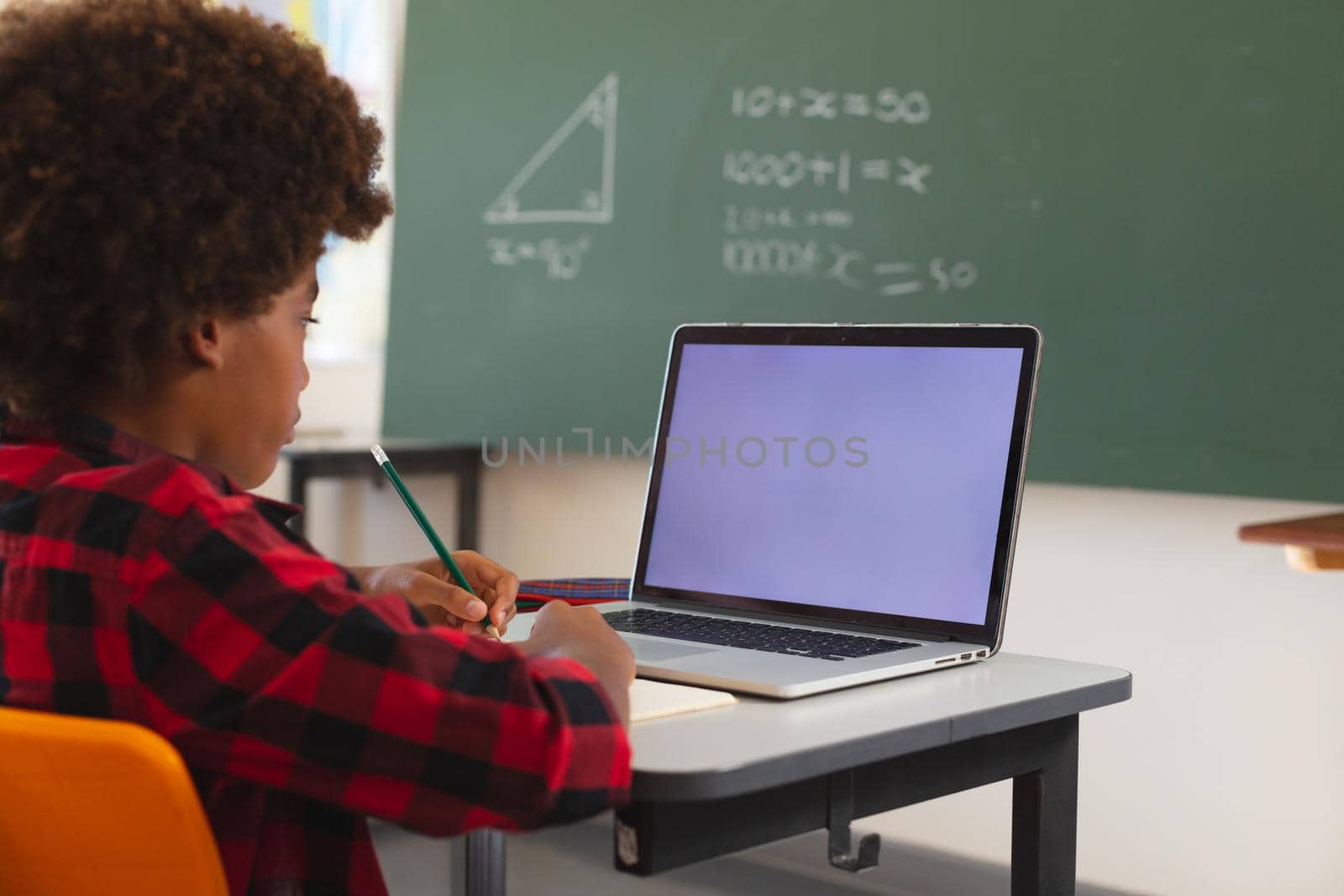 African american schoolboy sitting at desk in classroom using laptop, with copy space on screen by Wavebreakmedia