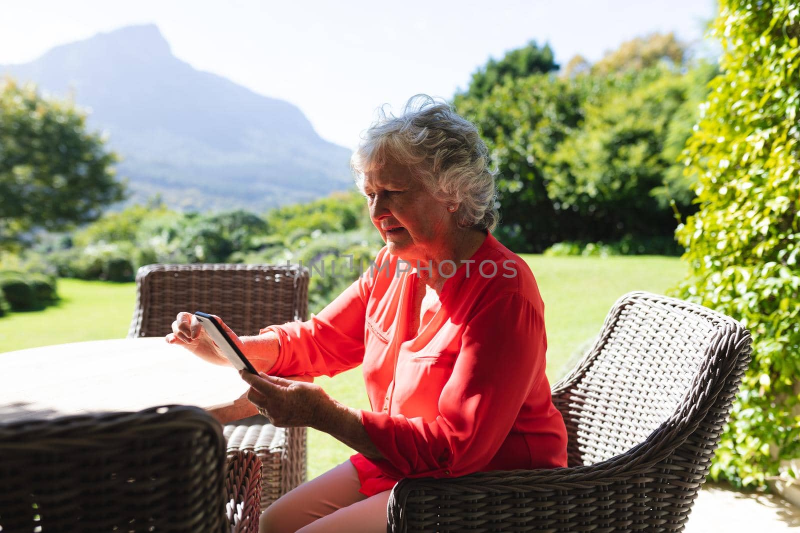 Senior caucasian woman sitting at table using smartphone in sunny garden. retreat, retirement and happy senior lifestyle concept.