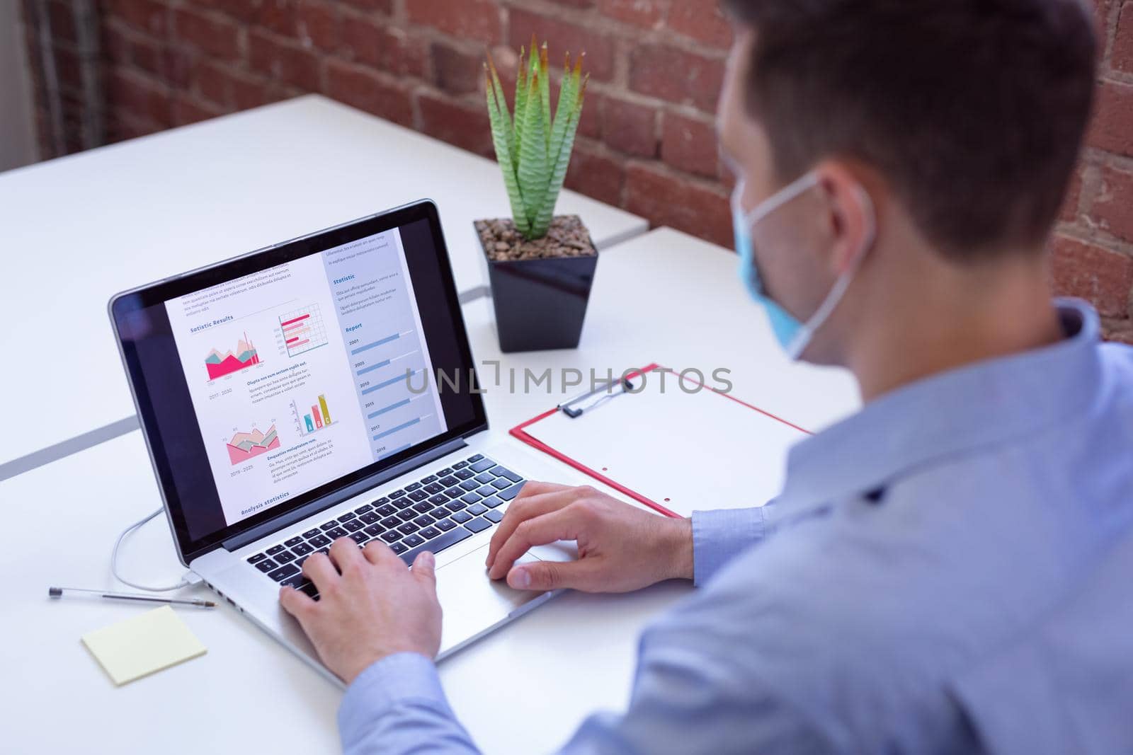 Caucasian businessman wearing face mask sitting at desk in office using laptop. working in business at a modern office during coronavirus covid 19 pandemic.